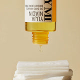 An upside-down bottle of Yuja Niacin Brightening Toner releases a drop onto stacked white cotton pads, set against a beige background.