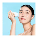 A woman with smooth, dewy skin applies the white rectangular Waterfull Hyaluronic Cooling Sunstick to her cheek. Her dark hair is pulled back and the light blue background enhances her fresh complexion.